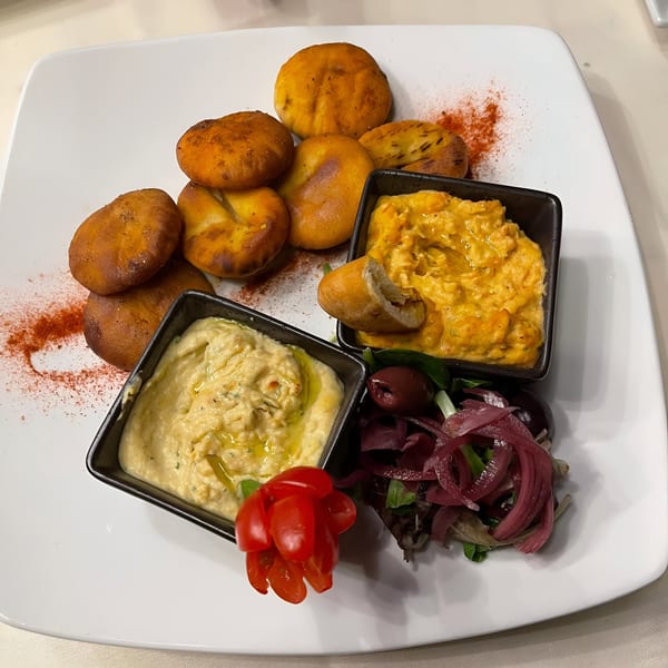 Mediterranean appetizer with pita bread, two dips, a small salad, and cherry tomatoes on a white plate.